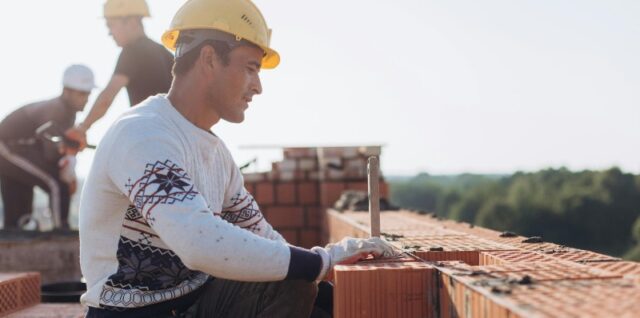 Construction worker laying bricks outdoors in sunny weather with lush greenery in background., tags: die - pexel